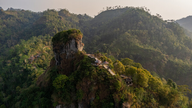 Aerial view of Puncak Widosari featuring its distinctive rocky peak, lush green hills, and small gazebos nestled in the landscape of Kecamatan Samigaluh, Daerah Istimewa Yogya, Indonesia.