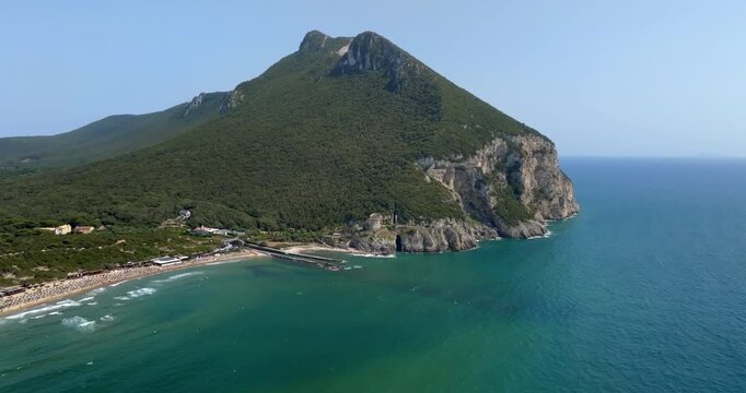 Aerial view of Mount Circeo, located in Lazio, Italy, rising above turquoise sea waters, with a historic watchtower perched on rocky coastal cliffs near a sandy beach stretch in a sunny summer morning