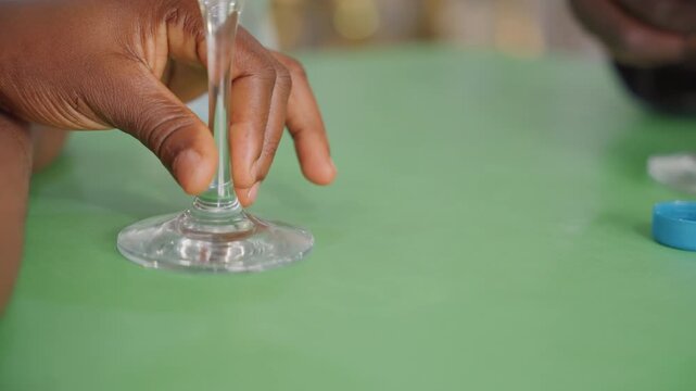 Black couple hands touch wine glass green, playful exchange near blue bottle cap on green table, two sets of fingers interacting in candid cafe scene, flirtatious gesture and shared moment, cozy