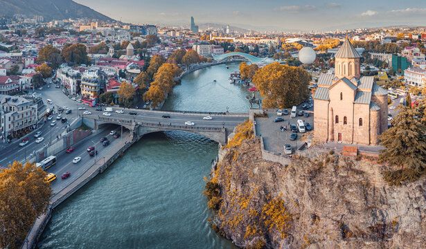 Metekhi Church on a cliff overlooking the Kura River and Tbilisi city skyline, featuring bridges and a cityscape at sunset