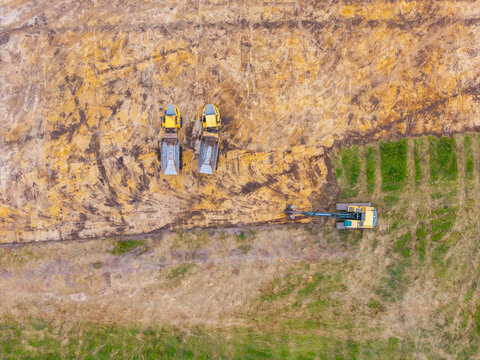 Aerial view of construction site with two yellow dump trucks and an excavator working on cleared soil surrounded by green grass in Valgunde Parish, Jelgava Municipality, Latvia.