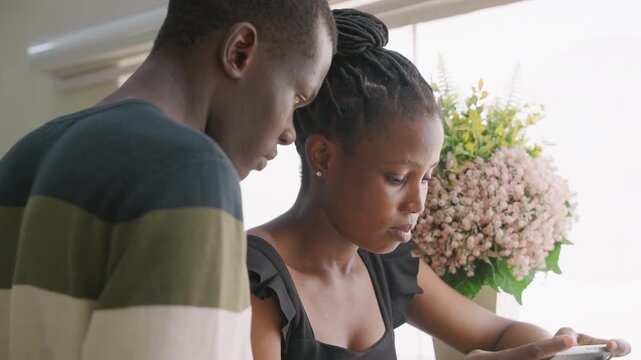 Black couple at kitchen counter studying phone together booking reservation, choosing restaurant, arranging surprise date with bouquet of flowers in background, soft daylight, tender focused