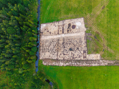 Aerial view of a rectangular patch of cleared soil bordered by a forest and a small stream in the middle of green agricultural fields in Valgunde Parish, Jelgava Municipality, Latvia.