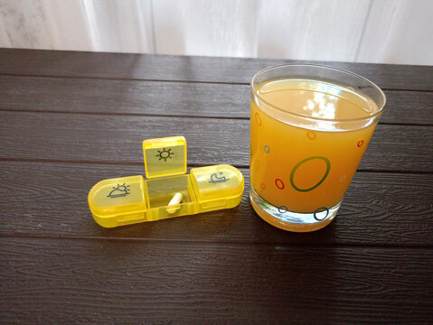 An open medication container with a white iron capsule and a glass of orange juice on a table. Iron deficiency anemia treatment - horizontal photo, close-up, side view