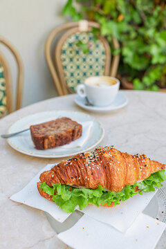 Croissant sandwich with fresh lettuce, banana bread, and coffee cup on a cafe table, enjoying a delightful breakfast outdoors