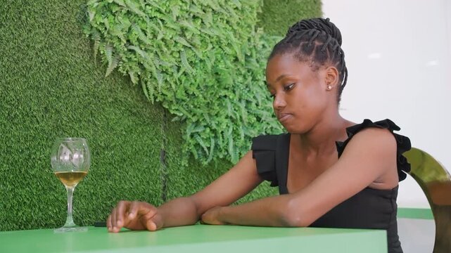 Woman sitting at green cafe table, gazing at wine glass and tapping fingers, empty chair opposite, indoor plant wall backdrop, soft natural light, subdued mood of disappointment and longing