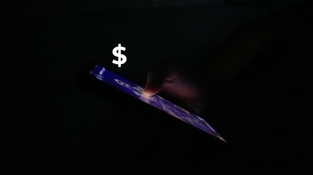 A hand holding a smartphone in the dark with a dollar sign icon.