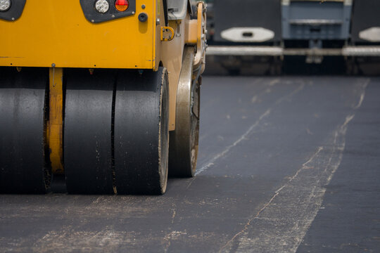 Close-up of yellow road roller compactor. Black drums press fresh asphalt. Smooth, dark pavement surface. Construction site backdrop. Professional machinery at work