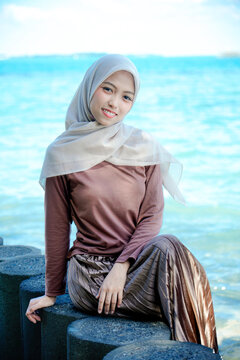 Medium three-quarter portrait of a lovely young Asian Muslim woman in a beige hijab and brown long-sleeved top sitting gracefully on concrete breakwater rocks.