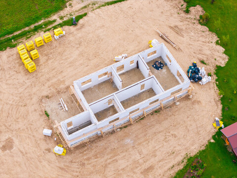 Aerial view of a house under construction with white block walls, stacks of yellow building blocks, and construction materials on a sandy lot, Jaunberze, Dobele Municipality, Latvia.