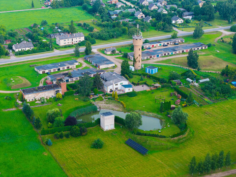 Aerial view of a rural landscape featuring a prominent water tower, a small pond, surrounding green fields, and residential buildings in Jaunberze, Dobele Municipality, Latvia.