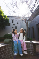 Fototapeta premium African American teenage females sitting and chatting on brick wall at campus courtyard with duffel