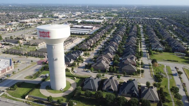 Concrete municipal water vessel towers beside residential streets near Phillips Creek Ranch. High angle drone shot highlights two story brick homes, dark roofs, paved driveways, Frisco, TX