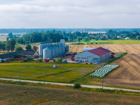 Aerial view of a farm complex with metal grain silos, a large storage shed, and hay bales in agricultural fields in Jaunberze Parish, Dobele Municipality, Latvia.