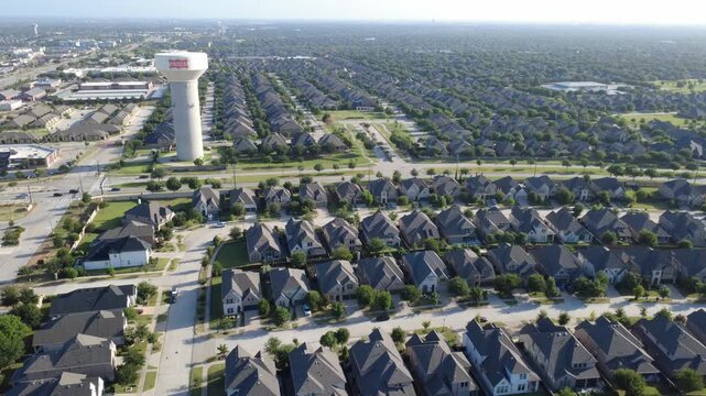 Plentiful single family home blocks flank local thoroughfares near Phillips Creek Ranch. Wide drone view highlights tree lined streets, manicured yards, municipal tower, Frisco, Texas