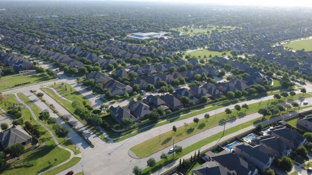 Manicured boulevard medians divide suburban housing tracts near Phillips Creek Ranch in North Texas. High aerial shot highlights moving vehicles, residential lawns, tree canopy, Frisco, TX