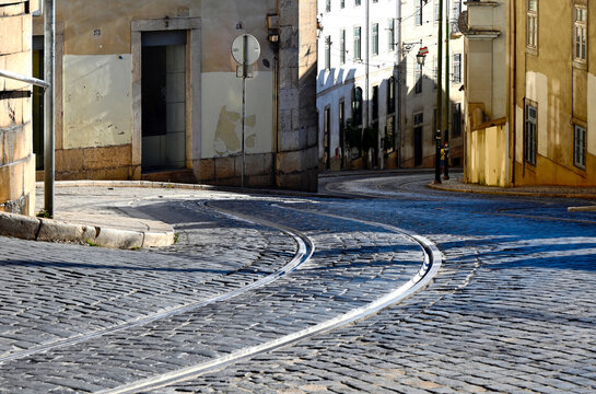 winding old street in Lisbon, Portugal. sloped cobble stone street pavement with steel streetcar rails. old stucco finished grunge residential buildings. car and pedestrian passing by. travel concept