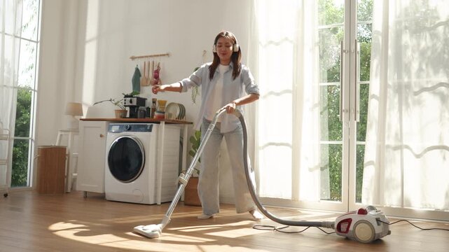 Happy young Asian woman cleaning floor with vacuum cleaner at home. Cheerful female enjoying domestic chores while listening to music and dancing energetically in modern room.