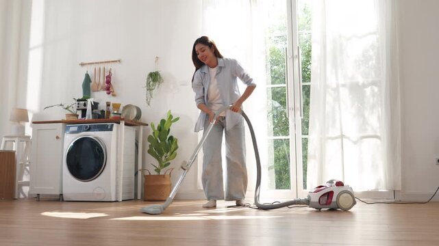 Happy young Asian woman cleaning floor with vacuum cleaner at home. Cheerful female enjoying domestic chores while listening to music and dancing energetically in modern room.