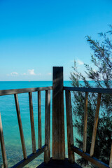 Fototapeta premium Scenic view of a rustic wooden balcony fence overlooking clear turquoise ocean water and green coastal pine trees under a bright blue sky. 