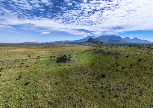 Aerial view of Pian Upe Game Reserve featuring vast grassy plains, scattered trees, and distant rugged mountains under a cloudy blue sky in Nakapiripirit, Northern Region, Uganda.