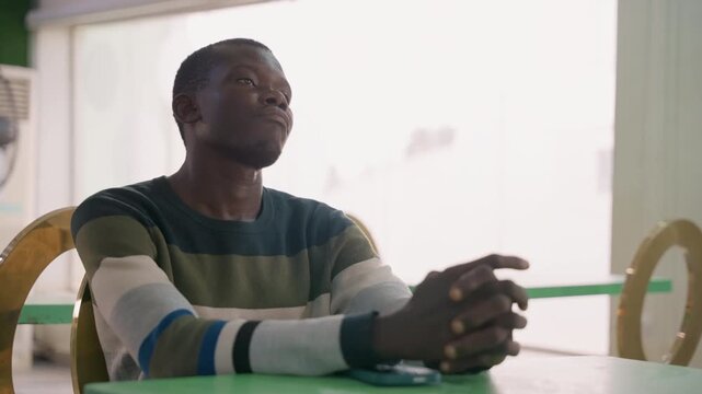 Seated black man waiting at table striped sweater hands clasped warm daylight pouring through window cozy cafe bistro setting hopeful expression nervous anticipation before romantic meeting
