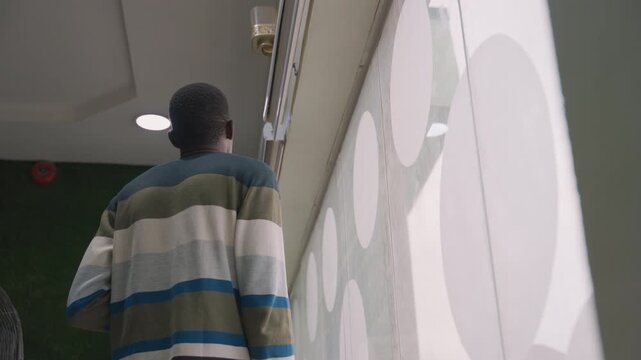 Black man peeking through patterned curtain. Glancing outside from modern interior, hopeful expression, adjusting blinds, soft overhead light, casual sweater, preparing to step out for date