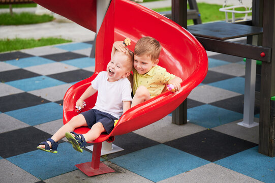 Two boys sliding together on red playground slide, laughing and holding toys, joyful outdoor play, park setting, happy childhood friendship moment