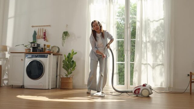 Happy young Asian woman cleaning floor with vacuum cleaner at home. Cheerful female enjoying domestic chores while listening to music and dancing energetically in modern room.
