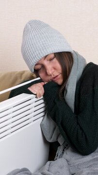 Cold woman wearing a beanie, sweater, and scarf trying to warm up by hugging a portable electric radiator during winter, illustrating the global energy crisis and heating costs