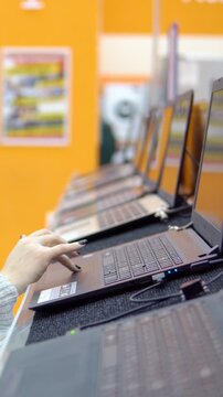 Female customer's hand with painted nails typing and scrolling on a new laptop in a computer store, browsing different models displayed in a row before making a purchase