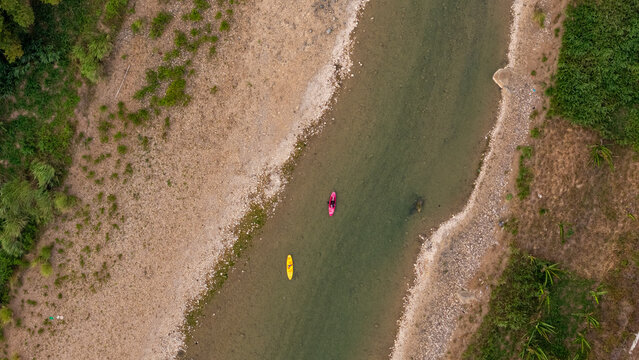 Aerial view of Sungai Oya with a pink kayak and a yellow kayak floating on the river, surrounded by sandy banks and lush vegetation in Kecamatan Imogiri, Daerah Istimewa Yogya, Indonesia.