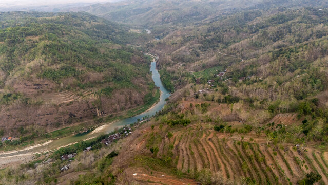 Aerial view of the winding Sungai Oya river flowing through lush green hills and terraced agricultural fields in Kecamatan Imogiri, Daerah Istimewa Yogya, Indonesia.