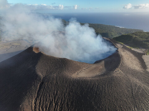 Aerial view of Mount Yasur volcano crater emitting thick white smoke with rugged volcanic slopes and lush green surroundings in Tafea Province, Vanuatu.