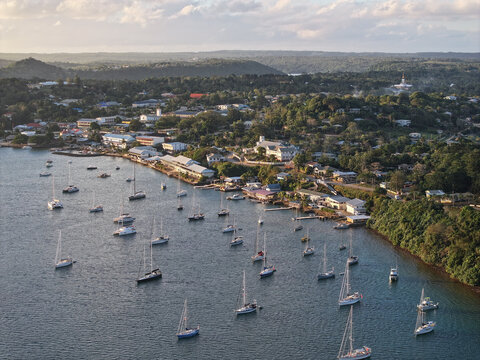 Aerial view of sailboats anchored in the bay, with lush green hills, coastal buildings, and calm waters under soft light Vava'u, Tonga.