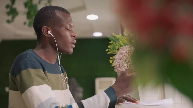 Black man listening to music while writing in cafe, young black creative with earbuds scribbling journal pages next to bouquet, relaxed posture, soft bokeh, green wall background, warm overhead