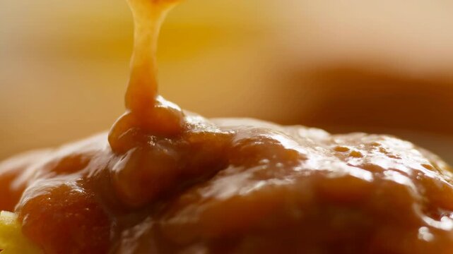 Close-up pouring orange sauce forming glossy bead over textured food surface under warm directional light with bubbles, reflections, and rich appetizing color, with sauce, pour, drip, closeup detail