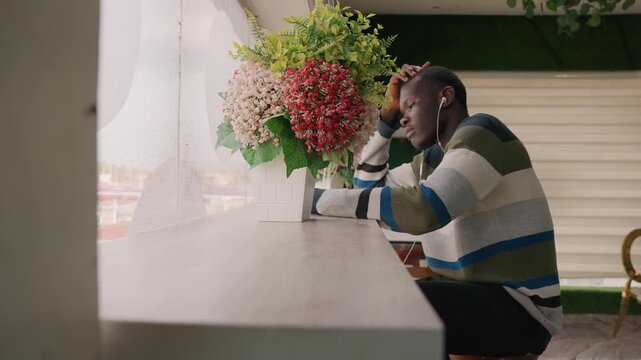 Black man sitting at cafe counter, gaze toward window beside large bouquet of flowers, wearing striped sweater, hand supporting head, anxious yet hopeful expression waiting for romantic date, soft