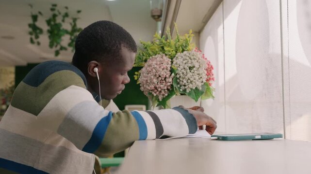 Black man at cafe table, solo date waiting while listening to music through earphones, striped sweater, tablet on table, bouquet near window, soft daylight, introspective mood, urban coffee shop