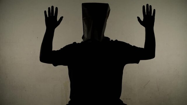 An anonymous silhouette of a man with a bag on his head stands against a dirty white wall with his hands raised