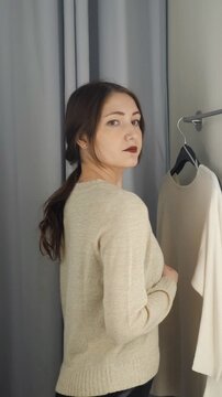 Beautiful young woman with dark lipstick trying on a new beige knitted sweater in a store's fitting room, looking at herself in the mirror with a satisfied smile