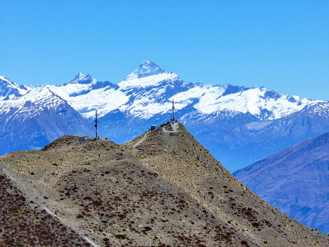 Aerial view of Roys Peak with communication towers on the ridge, dry grassy slopes, and snow-capped mountains in the background in Wanaka, Otago Region, New Zealand.