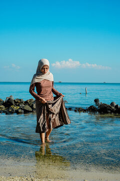 ​A young Asian Muslim woman in a hijab holding her skirt while standing in clear, shallow ocean water near a rocky shore under a bright blue sky. Concept of tropical vacation, modesty travel, peaceful