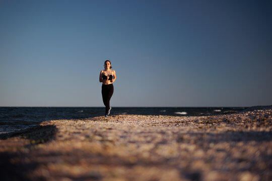 Fitness Woman Running On Sandy Beach Under the Blue Sky. Athletic girl enjoys outdoor cardio training near ocean waves during beautiful summer sunrise