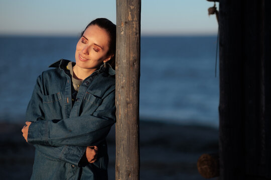 Melancholic Woman In Denim Jacket Leaning On a Post. Thoughtful female tourist with closed eyes rests near coast wearing autumn clothing during windy day
