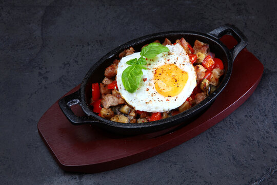 Black cast-iron mini frying pan with fried eggs, meat, pepper and mushrooms, decorated with basil, on red wooden tray and gray stone background