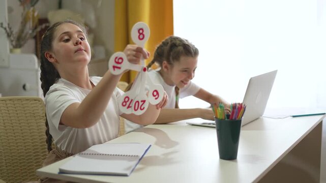 Young girl with braided hair holding math flashcards showing numbers while studying at home. Her sister works on laptop computer nearby. Notebooks and colorful pencils on white study table.