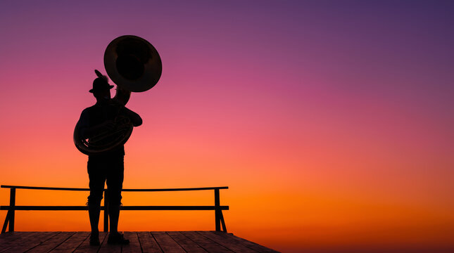 Silhouette of musician playing tuba brass instrument on wooden stage during vivid orange sunset. Live music festival outdoor background. Bavarian carnival Oktoberfest celebrating concept copy-space.
