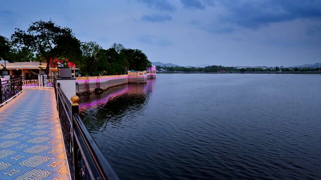 Bright decorative lighting illuminates patterned pathways near the water. Dark monsoon clouds hover overhead. Peaceful twilight fills Fateh Sagar Lake in Udaipur, Rajasthan.