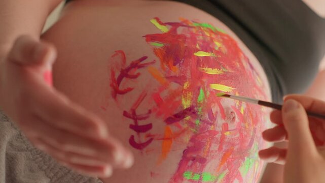 Pregnant caucasian belly painting brush strokes closeup, artist applies vivid neon pigment with careful gestural marks, cradling hand supports bump, bright colorful texture, joyful creative